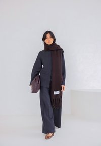 Chemise gris foncé à rayures fines et pantalon large, assortis à une longue écharpe marron, tenant un sac marron. Étiquette imprimée sur l'écharpe. Chaussures à imprimé léopard.