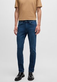 Dark blue denim jeans with a slim fit, featuring five-pocket styling and subtle fading. Paired with a light brown short-sleeve shirt.