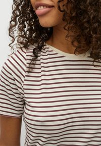 Camiseta de rayas en crema con rayas burdeos, hecha de tela suave. Presenta un cuello redondo y mangas cortas. Cabello rizado parcialmente visible.