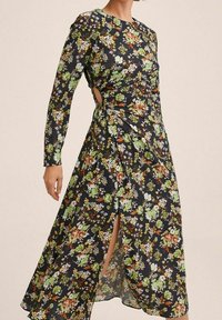Květinové šaty s dlouhými rukávy, černé pozadí, živé zelené, oranžové a bílé vzory, detail s bočním výřezem a rozparkem až na stehna.