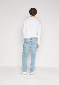 Vit långärmad skjorta, ljusblå denimjeans med en blekt struktur, och vita skor med svarta accenter. Sedd bakifrån.