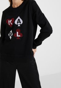 Svart sweatshirt med färgglada glittriga kortleksapplikationer: hjärta, spader och klöverdesigns. Har rund hals och ribbade muddar.