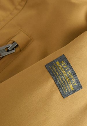 Braunfarbener Stoff mit glatter Textur und einem angenähten schwarzen Etikett. Das Etikett zeigt gelben Text, der Informationen zur Marke und zum Produkt enthält.