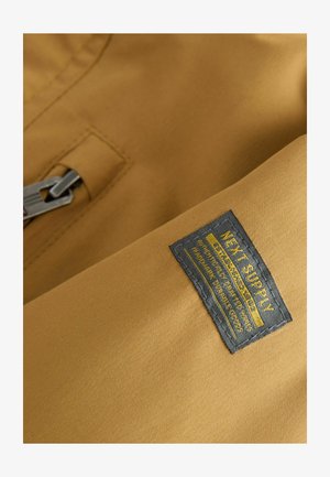 Tessuto beige che mostra una superficie liscia con un'etichetta nera cucita. L'etichetta presenta un testo giallo che specifica le informazioni sul marchio e sul prodotto.