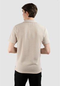 Ljusbeige kortärmad polo med ribbad textur, som har en mjuk krage och subtila vertikala ränder, tillsammans med mörka byxor.