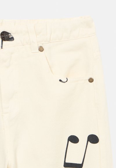 Beżowe spodnie z metalowym guzikiem i nitami, z czarnym motywem nuty muzycznej obok kieszeni na udzie.