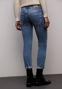 Smala jeans i ljusblå denim, med uppvikta benslut, bakfickor och en fyrkantig läderetikett vid midjan. Svarta anklar stövlar bärs.