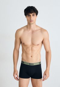 Schwarze Boxershorts mit passgenauer Passform und einem kontrasthaltigen khakifarbenen Logo-Bund, getragen von einem shirtlosen männlichen Model mit kurzen, lockigen Haaren.