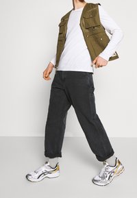 En person som bär en olivgrön arbetsväst, vit långärmad skjorta, svarta cargo-byxor, grå strumpor och sneakers med vitt och guldaccent stående med benen isär.