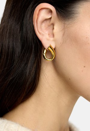 Ucho kobiety z grubym, błyszczącym złotym kolczykiem w kształcie skręconej pętli, ciemne włosy założone za ucho, jasny odcień skóry.