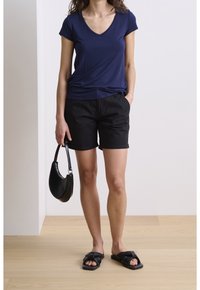 Camiseta navy de cuello en V, pantalones cortos negros con dobladillo enrollado, bolso negro y sandalias de tiras con detalle de nudos. Suelo de madera en un entorno neutro.