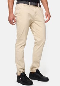 Pantalones beige de corte slim con bolsillos laterales, un cinturón tejido negro y zapatillas negras con logo. El material parece suave y ligero.