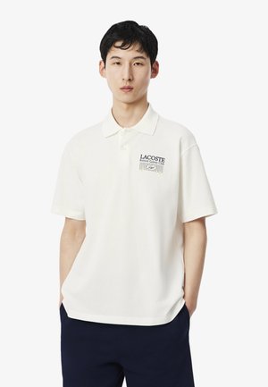 Joven con cabello corto y negro, vistiendo una camisa polo blanca de Lacoste con el logotipo del Club Rolands Garros y pantalones azul marino sobre fondo blanco.