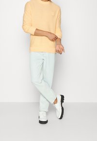 Man med persikofärgad tröja, ljusblå jeans och vita skor står med korslagda ben och justerar sin vänstra ärm mot en enfärgad bakgrund.