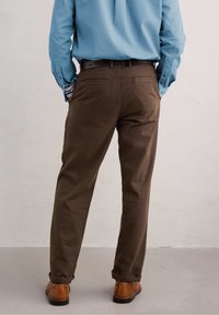 Hnědé kalhoty vyrobené z texturované látky, s bočními kapsami, přímým střihem a ohrnutými manžetami u kotníků, kombinované s hnědými botami.