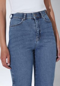 Jeans de mezclilla azul de cintura alta con cremallera, pasadores para cinturón y detalles de costura sutiles. Tinte medio con una textura suave.