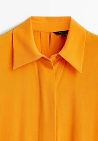 Kurzes oranges Blusenshirt mit klassischem Kragen, Knopfverschluss und Faltenverarbeitung an der Vorderseite. Glatte, leichte Stoffstruktur.