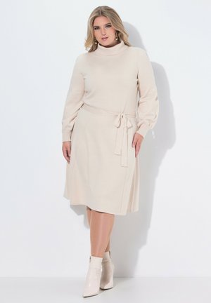Beige gebreide jurk met een col, lange mouwen en een strik in de taille. Glad textuur met een uitlopende rok. Gecombineerd met beige enkellaarsjes.