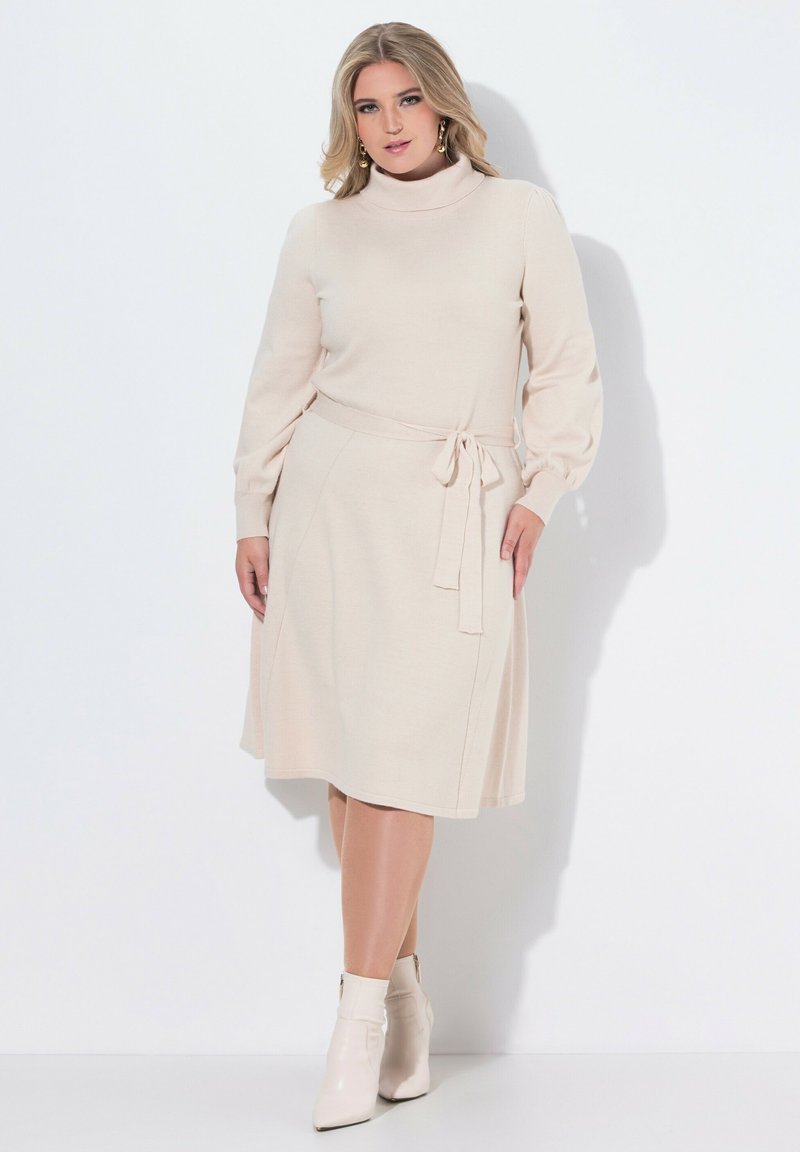 Abito in maglia beige con collo alto, maniche lunghe e un fiocco in vita. Texture liscia con gonna svasata. Abbinato a stivaletti beige alla caviglia.