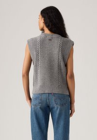 Grå strikket ermeløs genser med kabler langs sidene, ribbestrikket kant og rund hals, kombinert med blå denimjeans.