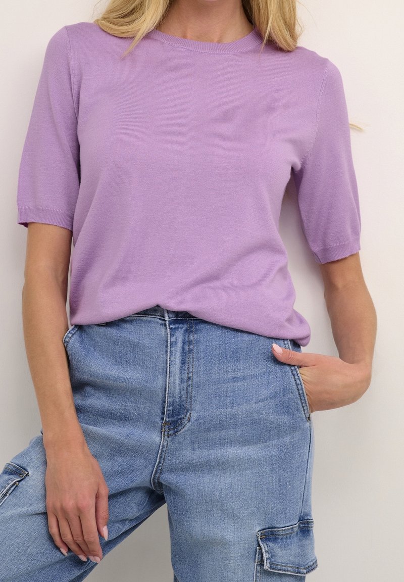 Femme portant un haut en maille lilas à manches courtes, rentré dans un pantalon cargo en denim bleu clair taille haute, une main dans la poche.