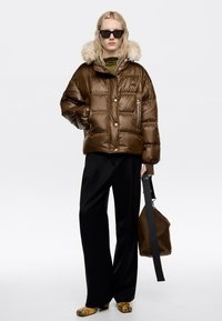 Hnedá pufovaná bunda s kožušinovou kapucňou, lesklý materiál, vrecko na zips, v kombinácii s čiernymi širokými nohavicami a hnedou taškou.