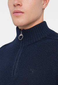 Maglione di lana navy con colletto alto a coste, dotato di chiusura zip e piccolo logo in rilievo sul lato sinistro del petto. Texture morbida.