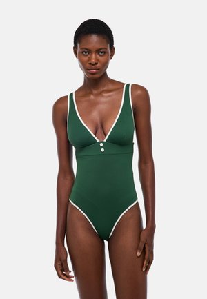 Femme portant un maillot de bain une pièce vert foncé avec un liseré blanc et deux boutons à l'avant, debout devant un fond uni.