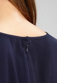 Námořnická modrá blůza s texturovanou tkaninou, ukazující zadní výstřih a malý černý zip uprostřed.