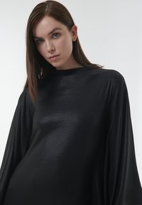 Zwarte langemouwen top met een zachte, glanzende textuur; wijde mouwen; hoge halslijn. De stof valt elegant, met een nadruk op een losse pasvorm.