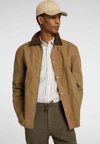 Chaqueta de algodón encerado marrón con cuello de pana, combinada con una camisa de rayas blanca y azules, gorra beis y pantalones verde oliva.