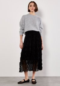 Maglione grigio lavorato a maglia con maniche lunghe, abbinato a una gonna midi nera, trasparente e voluminosa, e scarpe nere flat con cinturino alla caviglia.