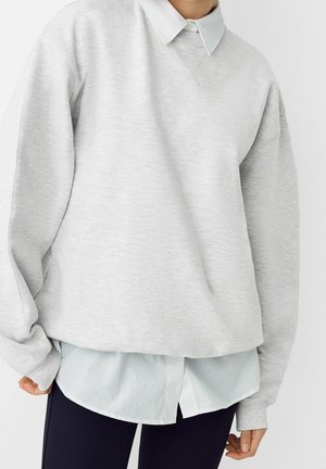 Persoon draagt een lichtgrijze trui over een wit overhemd met knopen en zwarte broek tegen een effen witte achtergrond.