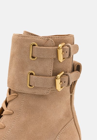Bottine en suède beige avec deux sangles à boucle dorée, fermeture à lacets sur le devant et une finition texturée sur le dessus.