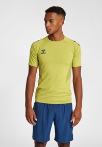 Jonge man in een limoen groene hummel sportshirt en blauwe sportshorts, staand en kijkend naar links tegen een witte achtergrond.