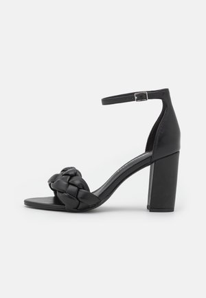 Sandale à talon noir avec une sangle tissée à l'avant, une sangle de cheville avec une boucle et un talon bloc épais. Texture en cuir lisse.