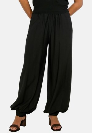 Vrouw draagt losse zwarte broek met elastische enkels, gecombineerd met donkerbruine open-toe sandalen met blokhak tegen een effen achtergrond.