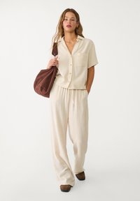 Béžový ľanový dvojdielny outfit pozostávajúci z krátkorečného košele na gombíky a voľných nohavíc s elastickým pásom. Hnedá kabelka cez rameno.
