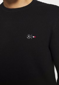 Maglione nero a coste con collo rotondo. Presenta il logo Mercedes ricamato e accenti colorati in rosso e bianco nella zona sinistra del petto.