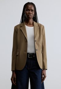 Beige skræddersyet blazer med hakkede revers, to knapper og forlommer, båret over en hvid skjorte og mørke denimbukser.