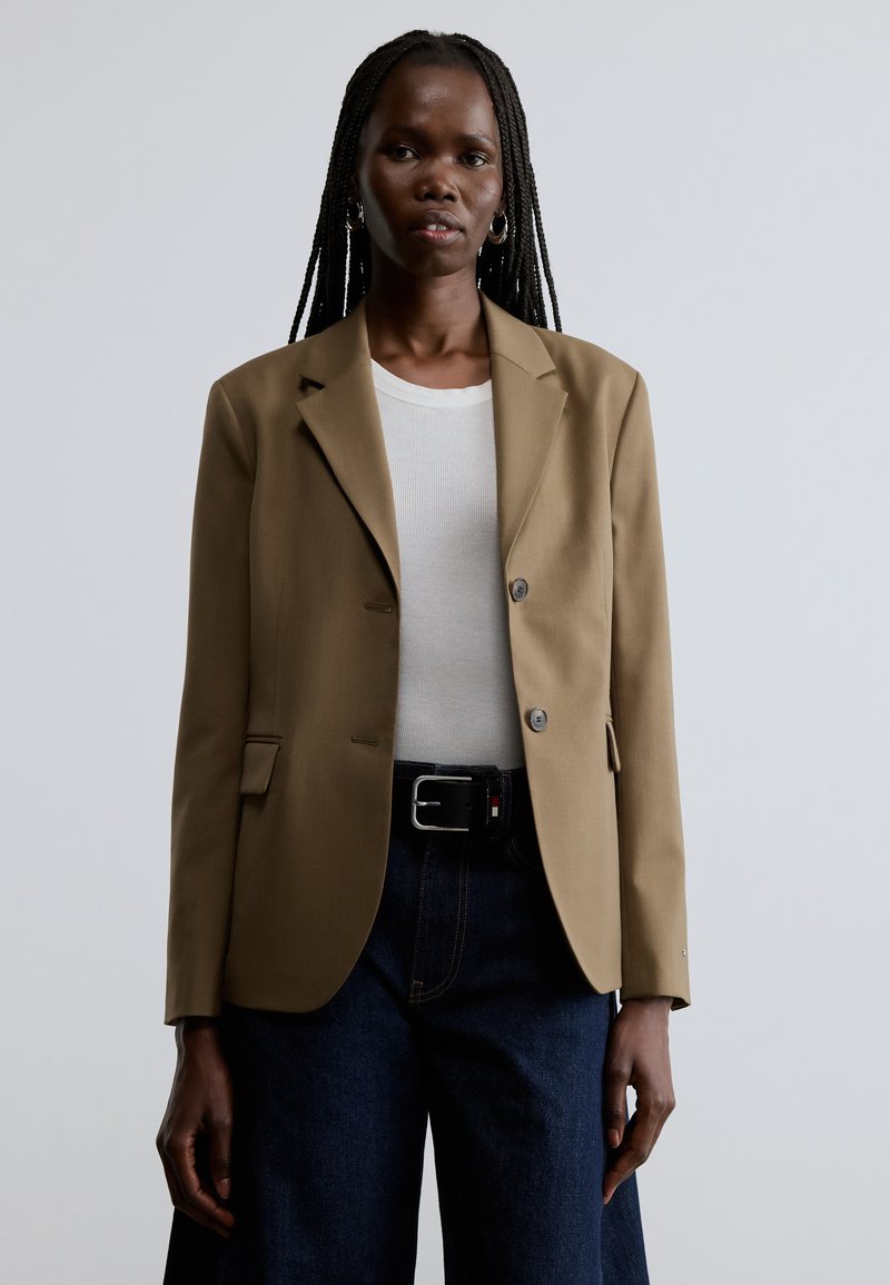 Beige skræddersyet blazer med hakkede revers, to knapper og forlommer, båret over en hvid skjorte og mørke denimbukser.