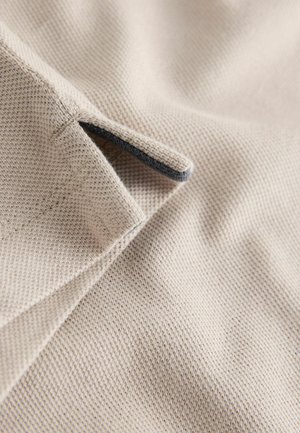 Primo piano di stoffa beige testurizzata con una piccola apertura e bordatura scura. Mostra un motivo intrecciato e una superficie liscia.