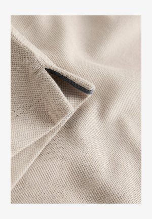 Bézs színű, texturált szövet közeli kép egy kis nyílással és sötét szegéllyel. Szövött mintázatot és sima felületet mutat.