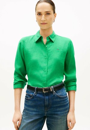 Mujer con una camisa verde de botones y mangas remangadas, jeans azules, cinturón negro y pequeños pendientes de aro, frente a un fondo blanco liso.