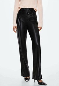 Femme portant un pantalon en simili cuir noir taille haute avec des coutures avant, associé à un pull rose clair et des escarpins noirs à bout pointu.