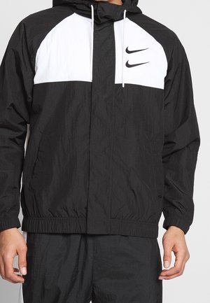 Giacca antivento bianca e nera con due loghi Nike Swoosh neri sul pannello bianco del petto, indossata con pantaloni neri coordinati.