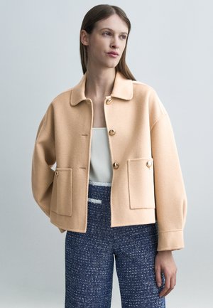 Kvinde iført beige uldjakke med knapper og store lommer, hvid top og blå teksturerede bukser, stående foran en ensartet lysegrå baggrund.