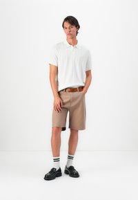 Young man standing with hands in pockets wearing white short-sleeve polo, beige shorts with brown belt, striped socks, and black loafers.