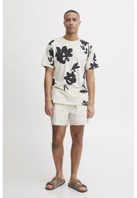 Short-sleeve beige t-shirt featuring large black floral prints, paired with light shorts and brown sandals, displaying a relaxed fit and casual style.