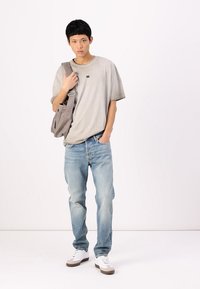 T-shirt gris à manches courtes avec un petit logo, assorti à un jean bleu clair et des baskets blanches avec des accents marron. Sac à bandoulière décontracté.